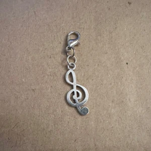 Silverfärgad berlock i form av en g-klav - Snygg och stilren berlock i form av en g-klav, perfekt för musikälskare! Den är silverfärgad och har en praktisk karbinhake för enkel fastsättning på armband eller halsband. Passar perfekt som en personlig touch till dina smycken eller som en present till någon speciell. 🎶