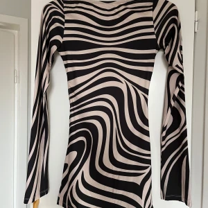 Svart och beige klänning med vågmönster - Snygg långärmad klänning med ett coolt svart och beige vågmönster. Den har en öppen rygg som ger en extra touch av stil. Perfekt för en utekväll eller fest!