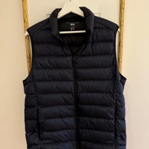  dunväst från Uniqlo - Säljer en stilren mörkblå dunväst från Uniqlo i storlek M. Västen är ultralätt och perfekt för lager-på-lager under höst och vår. Den har en dragkedja framtill och är i mycket bra skick. Perfekt för både vardag och utomhusaktiviteter.