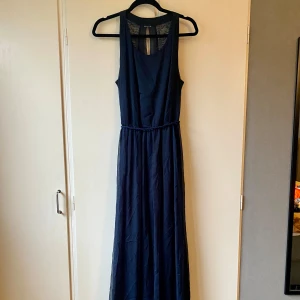 Mörkblå långklänning med spetsdetaljer - Säljer en elegant mörkblå långklänning med spetsdetaljer upptill. Klänningen är ärmlös och har en smickrande passform med ett knytband i midjan. Perfekt för festliga tillfällen eller en fin middag. Materialet är lätt och flödande, vilket ger en vacker siluett.