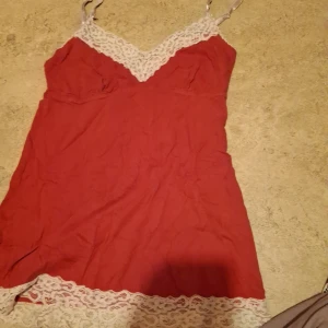 Röd linne med spetsdetaljer - Säljer en söt röd klänning med vita spetsdetaljer vid halsringningen och nederkanten. Klänningen är axelbandslös och har en kort längd, perfekt för sommarens fester. Materialet känns lätt och bekvämt. Passar bra till både vardag och festliga tillfällen!