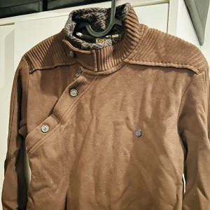 Brun tröja från LOCK - Säljer en brun tröja från LOCK i mycket bra skick. Den har en unik design med knappar på axeln och en hög krage. Perfekt för höst och vinter med sitt varma material. Tröjan är långärmad och har en snygg ribbad detalj vid axlarna.