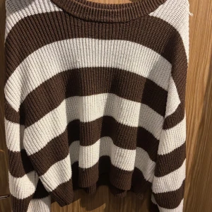 Brun och vit randig stickad tröja - Säljer en supermysig stickad tröja i brunt och vitt med breda ränder. Perfekt för höst och vinter, den är både varm och stilren. Tröjan har en avslappnad passform och långa ärmar, vilket gör den idealisk för kyliga dagar. Ett måste i garderoben för den som älskar bekväma och snygga plagg!