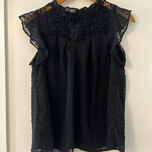 Svart ärmlös blus med spetsdetaljer - Snygg svart blus med spetsdetaljer och volanger vid ärmarna. Den är ärmlös och har ett diskret prickigt mönster. Perfekt för både vardag och festliga tillfällen. Blusen har en elegant och luftig design. Den är använd en gång hemma och är där av i nyskick. 