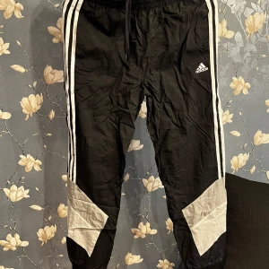 Svarta träningsbyxor från Adidas - Säljer ett par svarta träningsbyxor från Adidas med klassiska vita ränder längs sidorna. Byxorna har en elastisk midja med snörning och vita detaljer vid knäna. Perfekta för träning eller en avslappnad dag. De är i bra skick och redo för nya äventyr!