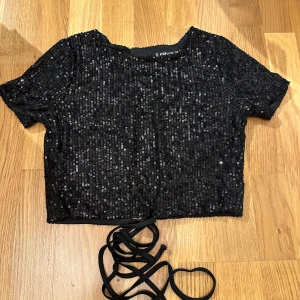 Svart glittrig topp med snörning - Säljer en superfin svart glittrig topp med paljetter. Den har korta ärmar och en öppen rygg med snörning som ger en snygg detalj. Perfekt för festliga tillfällen när du vill glänsa lite extra! ✨