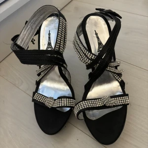 Svarta klackskor med strassdetaljer - Snygga svarta klackskor med glittriga strassdetaljer och en elegant rosett på sidan. De har en smal klack och flera remmar som korsar över foten för en chic look. Perfekta för festliga tillfällen när du vill glänsa lite extra! 🖤✨