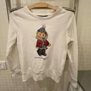 Vit tröja med björnmotiv från Ralph Lauren - Säljer en superfin vit tröja från Ralph Lauren med ett gulligt björnmotiv på framsidan. Tröjan har långa ärmar och är perfekt för en avslappnad stil. Den är i mycket bra skick och passar perfekt för höst och vinter.