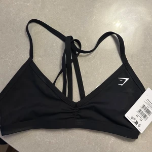 Svart sporttopp från Gymshark - Säljer en helt ny svart sporttopp från Gymshark. Den har en stilren design med smala axelband och en snygg detalj i ryggen. Perfekt för träning eller en aktiv vardag. Materialet är mjukt och stretchigt för bästa komfort.