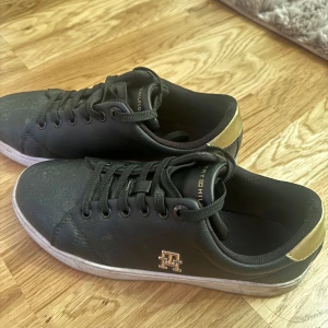 Svarta sneakers från Tommy Hilfiger - Säljer ett par svarta sneakers från Tommy Hilfiger i bra skick. De har snygga gulddetaljer vid hälen och loggan på sidan. Skorna har snörning och en vit sula som ger en stilren look. Perfekta för både vardag och fest!