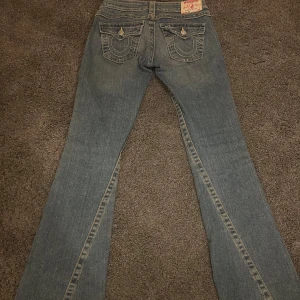Bootcut True Religion jeans  - Säljer ett par snygga blå jeans från True Religion i bra skick. Storlek: 26❤️ De har en bootcut-stil och är lågmidjade med coola detaljer på bakfickorna. Pris kan diskuteras🔥Midja rakt över: ca 36cm❤️innerbenslängd: ca 83-84cm❤️Ytterbenslängd: ca 103cm