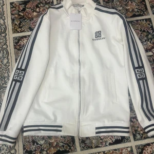 Vit jacka från Givenchy - Säljer en snygg vit jacka från Givenchy med svarta detaljer. Jackan har dragkedja framtill och långa ärmar med coola ränder längs sidorna. Perfekt för en stilren look, oavsett om du ska på fest eller bara hänga med kompisar. Givenchy-loggan syns både på bröstet och ärmarna. Passar perfekt till både vardag och lite finare tillfällen!