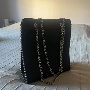 Svart axelväska med kedjedetaljer - Snygg svart axelväska i skinn med coola kedjedetaljer längs kanterna och som axelrem. Perfekt för en kväll ute eller en dag på stan. Väskan har en stilren design som passar till det mesta. Ett måste för garderoben!