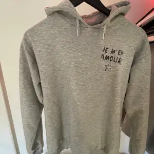 Säljer en grå hoodie från Gina Tricot med texten 'Je m'en amour' och en stjärna tryckt på både fram- och baksidan. Den är supermjuk och perfekt för en avslappnad stil. Passar perfekt till höst och vår. Hör av dig om du är intresserad! Den är lite sliten men när ja har haft på mig den så har ja aldrig tänkt på det