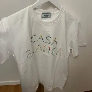 Säljer en superfin vit t-shirt från Casablanca med blommigt mönster som bildar texten 'CASABLANCA' på framsidan. Perfekt för en avslappnad och stilren look. T-shirten är i nytt skick och kommer i originalförpackning. Perfekt för sommaren eller våren! 🌼