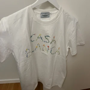 Vit t-shirt från Casablanca - Säljer en superfin vit t-shirt från Casablanca med blommigt mönster som bildar texten 'CASABLANCA' på framsidan. Perfekt för en avslappnad och stilren look. T-shirten är i nytt skick och kommer i originalförpackning. Perfekt för sommaren eller våren! 🌼