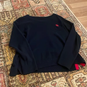 Mörkblå tröja från Abercrombie & Fitch - Säljer en snygg mörkblå tröja från Abercrombie & Fitch med röd logga på bröstet. Tröjan har långa ärmar och ribbade kanter. Perfekt för en avslappnad stil eller en kylig dag. Passar både till vardags och lite finare tillfällen.