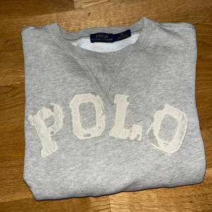 Grå sweatshirt från Ralph Lauren - Säljer en snygg grå sweatshirt från Ralph Lauren med texten 'POLO' i en cool, sliten stil på framsidan. Tröjan är i bra skick och perfekt för en avslappnad look. Den har långa ärmar och en klassisk rund halsringning. Perfekt för höst och vår!