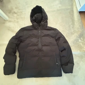 Svart pufferjacka från Jack & Jones - Säljer en svart pufferjacka från Jack & Jones i storlek L. Den är perfekt för kalla dagar med sin tjocka vaddering och huva. Jackan har en dragkedja framtill och är i mycket bra skick. Perfekt för vintern! ❄️