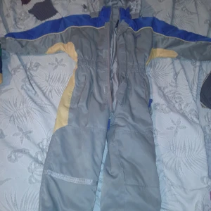 Grå och blå overall - Säljer en grå overall med blå och gula detaljer. Perfekt för utomhusaktiviteter under kalla dagar. Den har en dragkedja framtill och långa ärmar. Overallen är i bra skick och redo för nya äventyr!