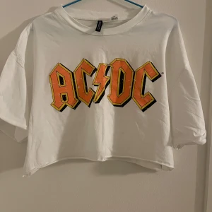 Vit AC/DC t-shirt - Säljer en cool vit AC/DC t-shirt från H&M. Den är croppad och har en stor, färgglad logga på framsidan. Perfekt för en avslappnad stil eller konsert! 🖤⚡