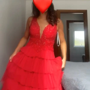 Röd balklänning med spetsdetaljer - Säljer en fantastisk röd balklänning med spetsdetaljer och volanger. Klänningen har en djup v-ringning och är ärmlös, perfekt för festliga tillfällen. Den har en vacker, flerdelad kjol som ger en elegant look. Perfekt för bal eller nyår!