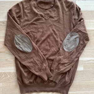 Brun tröja från Massimo Dutti - Säljer en snygg brun tröja från Massimo Dutti. Den har en v-ringad design och stiliga armbågslappar i en ljusare nyans. Perfekt för höst och vinter, både mysig och elegant. Passar bra till både vardag och lite finare tillfällen.