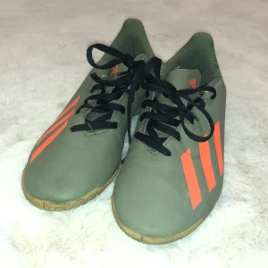 Gröna Adidas fotbollsskor med orange detaljer - Säljer ett par gröna Adidas fotbollsskor i storlek 36. De har snygga orangea ränder och svart snörning. Perfekta för inomhusfotboll eller träning. Skorna är i bra skick och redo för nya äventyr på planen!