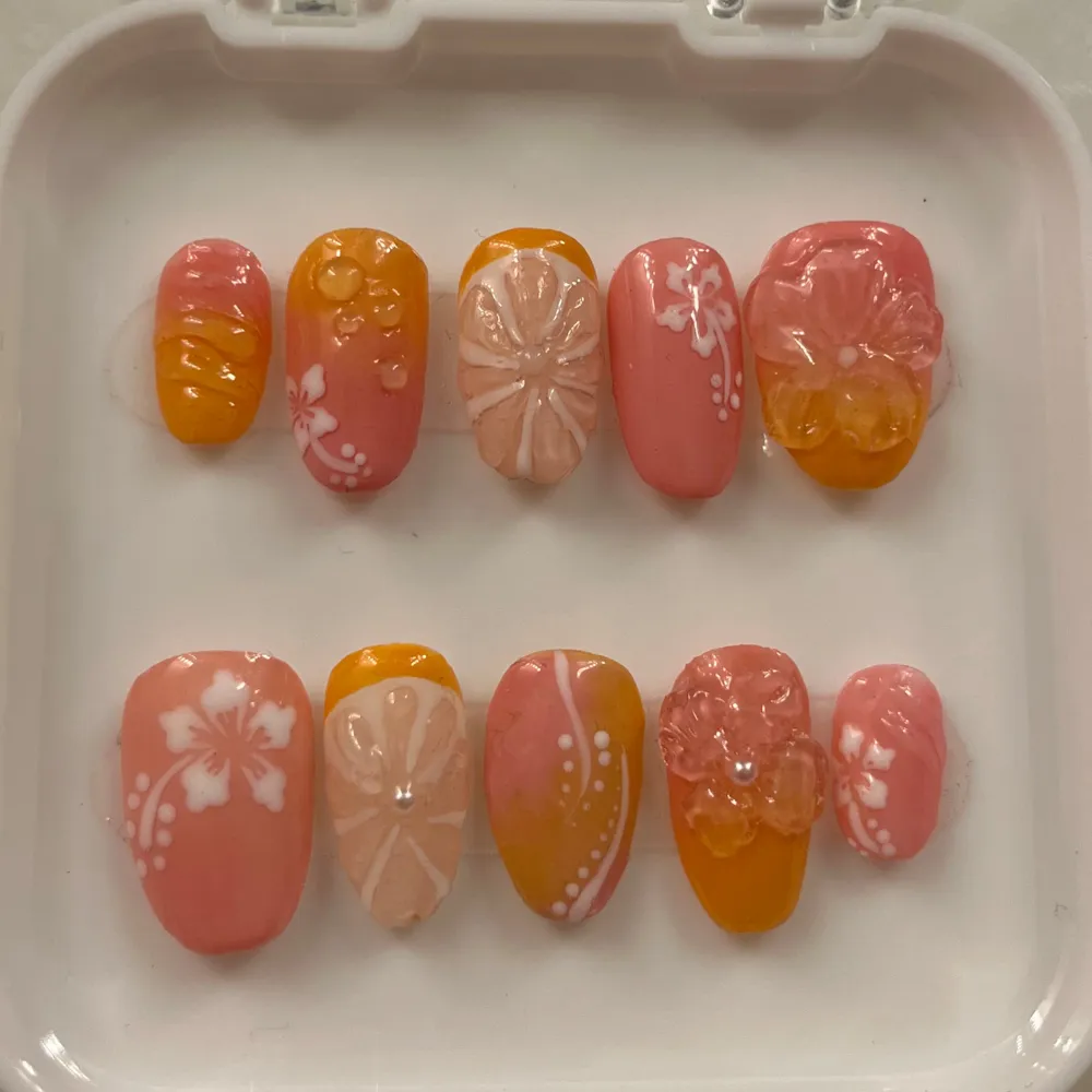 Snygga lösnaglar i rosa och orange med blommönster och 3D-blommor. Perfekt för en somrig look eller en festlig tillställning. Naglarna har en glansig finish och detaljer i vitt som ger en extra touch av elegans.. Asusteet.