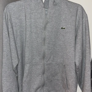 Grå hoodie från Lacoste - Säljer en klassisk grå hoodie från Lacoste i storlek L. Men passar även M. Den har en dragkedja framtill och den ikoniska krokodilloggan på bröstet. Perfekt för en avslappnad stil och passar till alla säsonger. Superbekväm och stilren!