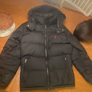 Säljer en snygg svart dunjacka från Ralph Lauren. Den har en huva och dragkedja framtill, perfekt för kalla dagar. Jackan är i mycket bra skick och passar både höst och vinter. Som ni ser på sista bilden är den där grejen sönder tyvärr. Är 183 cm lång skulle säga att passar 170-183 bäst. Storlek 14-16 vilket ungefär motsvarar xs-s i vanlig storlek