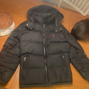 Svart dunjacka från Ralph Lauren - Säljer en snygg svart dunjacka från Ralph Lauren. Den har en huva och dragkedja framtill, perfekt för kalla dagar. Jackan är i mycket bra skick och passar både höst och vinter. Som ni ser på sista bilden är den där grejen sönder tyvärr. Är 183 cm lång skulle säga att passar 170-183 bäst. Storlek 14-16 vilket ungefär motsvarar xs-s i vanlig storlek