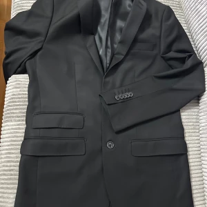 Svart kavaj - Säljer en stilren svart kavaj i mycket bra skick. Den har en klassisk design med två fickor fram och en enkel knappstängning. Perfekt för både formella och semi-formella tillfällen. Passar bra till både byxor och jeans för en mer avslappnad look.