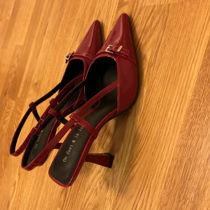 Röda pumps med spetsig tå - Snygga röda pumps i lack med spetsig tå och liten klack. De har en elegant rem med guldfärgat spänne över foten. Perfekta för en festlig kväll eller en utekväll med vänner. Skorna är i storlek 38 och ser ut att vara i mycket bra skick.