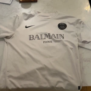 Vit fotbollströja från Nike x Balmain - Säljer en snygg vit fotbollströja från Nike i samarbete med Balmain. Tröjan har korta ärmar och ett stilrent tryck med Balmain Paris-loggan på bröstet. Perfekt för både träning och vardag. Passa på att köpa denna unika tröja!