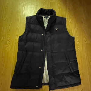 Navy dunväst från GANT - Säljer en snygg navy dunväst från GANT i mycket bra skick. Västen har en klassisk design med knappar och en randig insida. Perfekt för höst och vår när du vill hålla dig varm men ändå se stilren ut. Passar bra till både vardag och lite finare tillfällen.