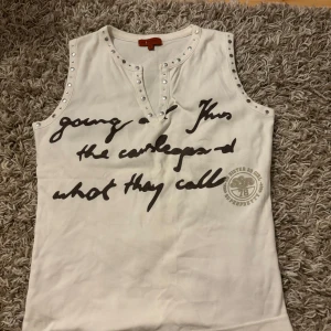 Vit ärmlös topp med text och nitar - Snygg vit ärmlös topp med cool text på framsidan. Den har nitar längs halsringningen och ärmarna för en edgy look.