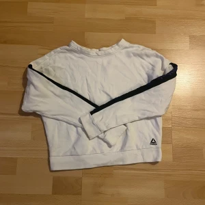 Vit tröja från Reebok med blåa detaljer - Säljer en snygg vit tröja från Reebok med blåa ränder längs ärmarna och Reebok-logga på sidan. Perfekt för en sportig look eller en avslappnad dag. Tröjan har långa ärmar och är i mycket bra skick. Passar perfekt till jeans eller träningsbyxor!