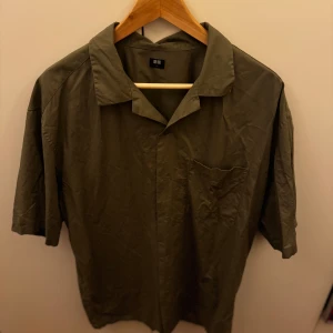 Olivgrön kortärmad skjorta från Uniqlo - Säljer en olivgrön kortärmad skjorta från Uniqlo i storlek L. Skjortan har en klassisk krage och en bröstficka. Perfekt för en avslappnad sommarlook eller en stilren vardagsoutfit. Materialet känns lätt och bekvämt. Passar både till jeans och shorts!