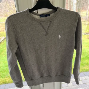 Grå sweatshirt från Polo Ralph Lauren - Säljer en stilren grå sweatshirt från Polo Ralph Lauren. Den har en klassisk rund halsringning och det ikoniska vita logotypbroderiet på bröstet. Perfekt för en avslappnad look och passar till alla säsonger. 