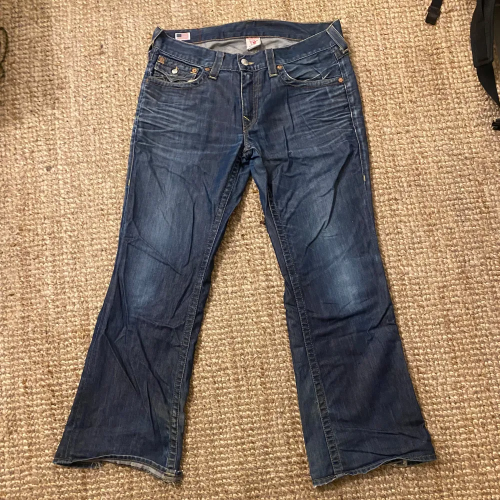 Säljer ett par blå jeans från True Religion i bra skick. De har en bootcut-stil och är tillverkade i USA. Jeansen har snygga bakfickor med knappar och slitna detaljer vid bensluten. Perfekta för en avslappnad look!. Jeans & Byxor.