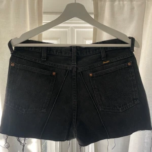 Svarta jeansshorts från Wrangler - Säljer ett par snygga svarta jeansshorts från Wrangler. De har en klassisk femficksdesign och råa kanter för en cool look. Perfekta för sommardagar eller festivaler. Passar till allt från t-shirts till blusar!