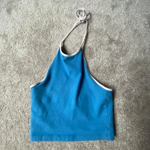 Blå halterneck-topp - Säljer en snygg blå halterneck-topp med vita kanter. Perfekt för sommardagar eller en avslappnad fest. Toppen är ärmlös och har en enkel knytning i nacken. Materialet känns mjukt och bekvämt.