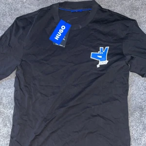 Svart t-shirt från Hugo - Säljer en svart t-shirt från Hugo i nytt skick. Den har en cool blå och vit detalj på bröstet som ger en unik touch. Perfekt för vardagsbruk eller en avslappnad utekväll. T-shirten är kortärmad och gjord i mjukt bomullsmaterial för bästa komfort.