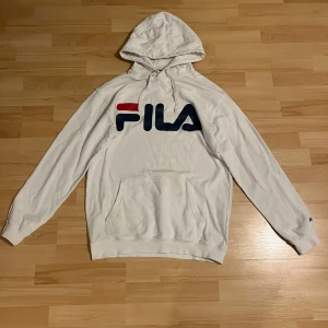 Herrhoodie - Säljer en snygg vit hoodie från FILA i storlek M. Den har en stor logga på bröstet i rött och blått, samt en praktisk magficka. Perfekt för en avslappnad stil och passar bra till både höst och vår. Huvan är justerbar med snören. Skön och bekväm att ha på sig!