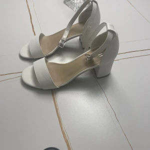 Vita klackskor  - Snygga vita klackskor i  med öppen tå och justerbart spänne runt ankeln. Endast använda en gång. Med lappen kvar. Köpt för 350kr.