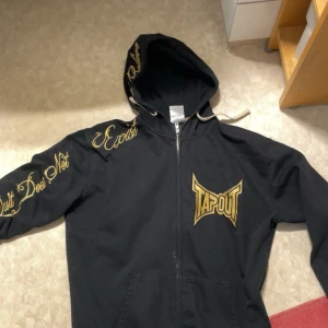 Svart hoodie från Tapout - Säljer en svart hoodie från Tapout med gulddetaljer. Den har texten 'Believe' på ryggen och 'Quit Does Not Exist' på ärmen. Bra kvalitet 