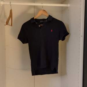 Säljer en stilren svart pikétröja från Ralph Lauren i slim fit. Den har en klassisk krage och korta ärmar, perfekt för både vardag och fest. Den röda loggan på bröstet ger en snygg kontrast. Passar perfekt till jeans eller chinos! Säljer pga att den är för liten för mig nu, inte använd så mycket, kan diskutera om priset eller byten 