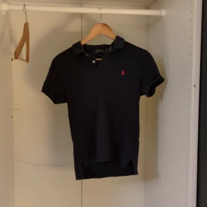 Mörkblå  pikétröja från Ralph Lauren - Säljer en stilren svart pikétröja från Ralph Lauren i slim fit. Den har en klassisk krage och korta ärmar, perfekt för både vardag och fest. Den röda loggan på bröstet ger en snygg kontrast. Passar perfekt till jeans eller chinos! Säljer pga att den är för liten för mig nu, inte använd så mycket, kan diskutera om priset eller byten 