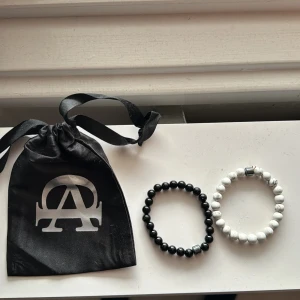 Allcapone pärlarmband  - Två stilrena armband med pärlor, ett i svart och ett i vitt. Perfekta för att matcha olika outfits. De kommer i en snygg svart påse med ett silverfärgat tryck. Armbanden är elastiska och passar de flesta handleder. Perfekt accessoar för både vardag och fest!